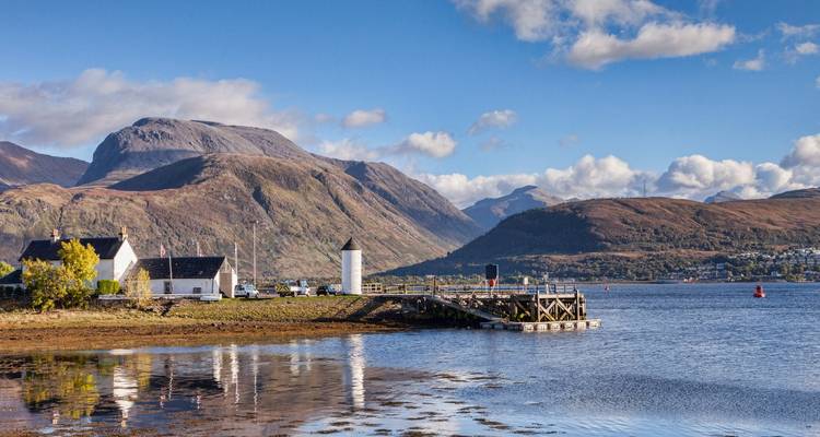 Ben Nevis elevándose sobre casitas costeras y un pequeño faro reflejado en las tranquilas aguas del loch.