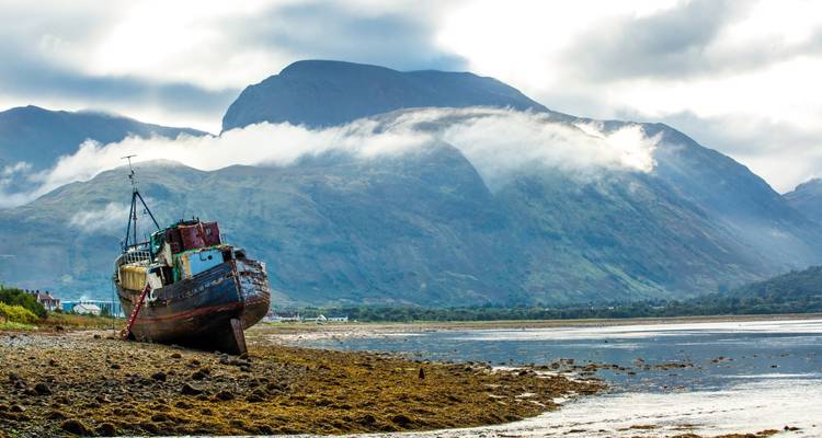 An abandoned fishing boat rests on a pebbled shore with mist-cloaked mountains towering behind.
