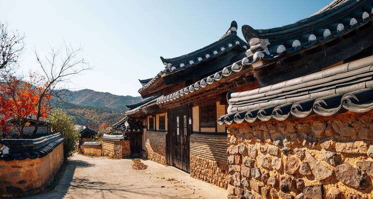 Traditional stone-and-wood Korean hanok with tiled roof set against forested hills and autumn leaves under clear skies.