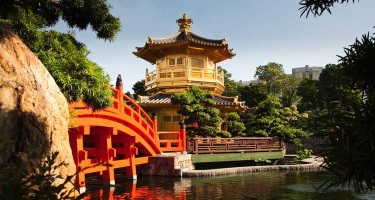 Golden octagonal pavilion and bright red arched bridge reflected in a tranquil garden pond beneath blue skies.