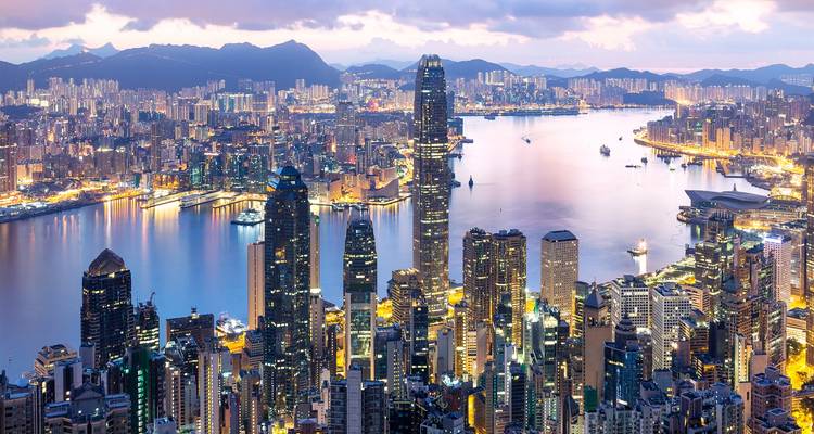 Sweeping twilight panorama of Hong Kong’s dense skyscrapers flanking Victoria Harbour with mountains beyond.