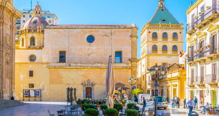 Sunny town square with ochre buildings, small café tables and people strolling in Marsala.