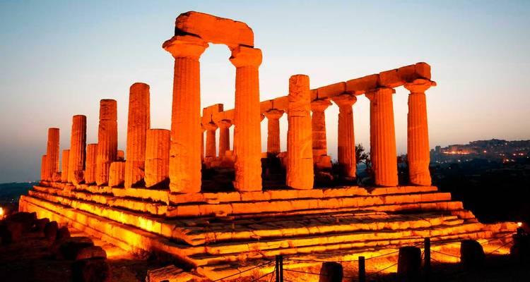 Dramatisch beleuchtete griechische Tempelruinen, die orangefarben gegen den Dämmerungshimmel leuchten.