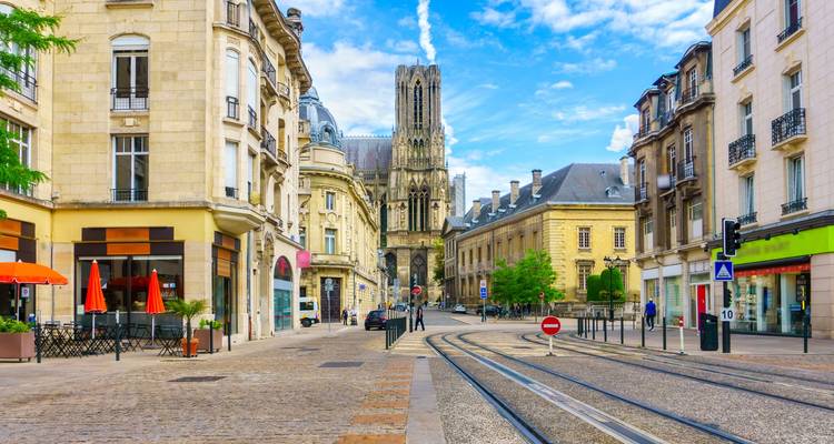 Calle con vías de tranvía que conducen a la Catedral de Reims rodeada de ornamentada arquitectura francesa en un día soleado.
