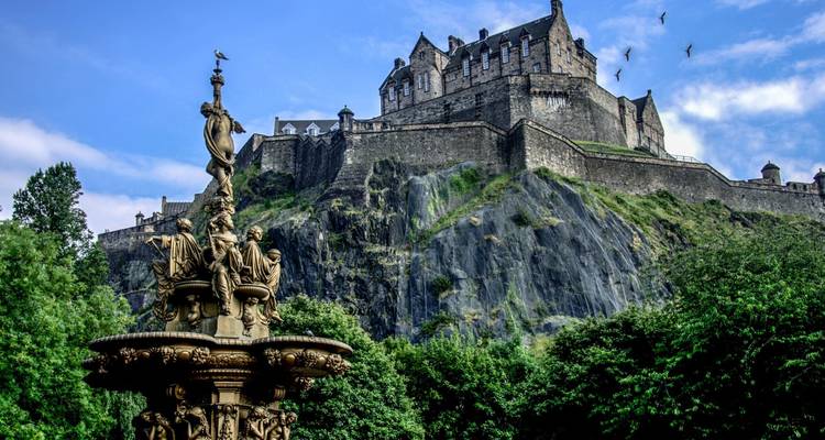 Ross Fountain in the foreground with Edinburgh Castle perched atop its rocky crag under a bright blue sky with birds soaring.