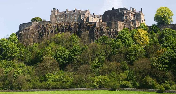 Stirling Castle sits atop a wooded cliff surrounded by green fields under bright spring sunshine.