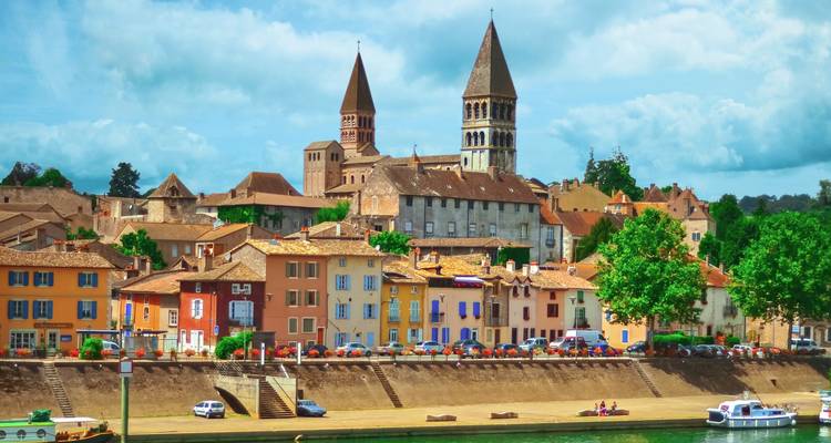 Riverside town of Tournus displays pastel houses and twin Romanesque church towers under fluffy clouds.