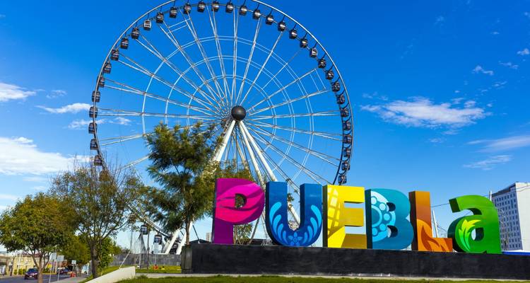 Panneau coloré PUEBLA devant une immense grande roue sous un ciel bleu éclatant.