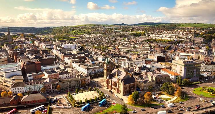 Aerial daylight view over the city of Cork with church spires, rolling green hills and scattered clouds.