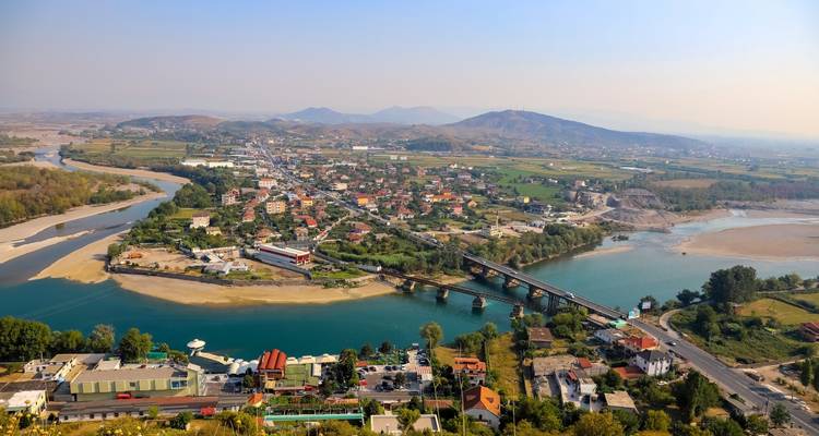 Wide aerial panorama of a river winding past bridges and a low-rise town in a green valley with hazy hills on the horizon.
