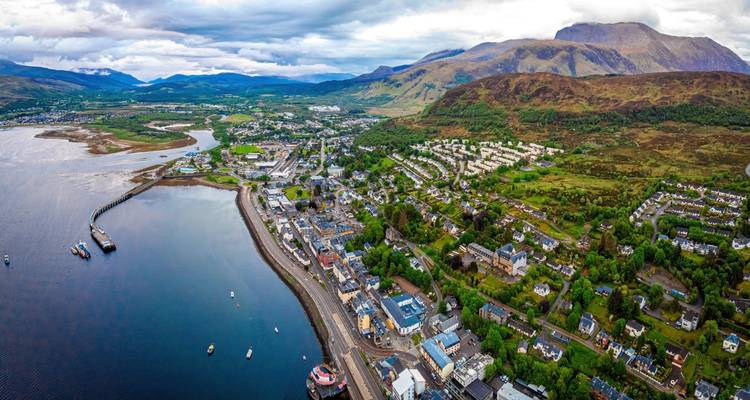 Vue aérienne large d'une ville côtière de montagne s'étendant le long d'un loch avec des montagnes en arrière-plan sous des nuages dramatiques.