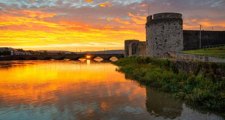 Le coucher de soleil flamboyant se reflète sur une rivière à côté de la tour en pierre médiévale et des murs d'un château au bord de la rivière.