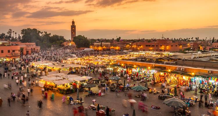 Place de marché animée en soirée remplie d'étals alimentaires et de foule sous un ciel au coucher de soleil orangé, tour de mosquée imposante en arrière-plan