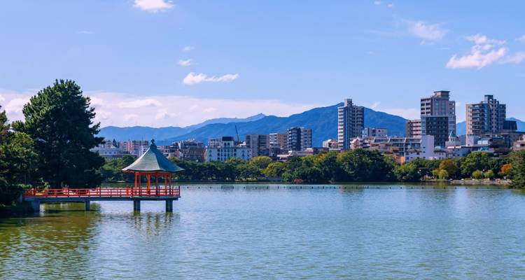 Serene lake with red-roofed pavilion and Fukuoka city skyline backed by distant mountains.
