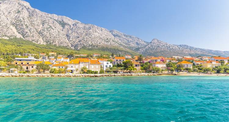 A quaint Adriatic seaside village with orange roofs sits below towering limestone mountains and turquoise water.
