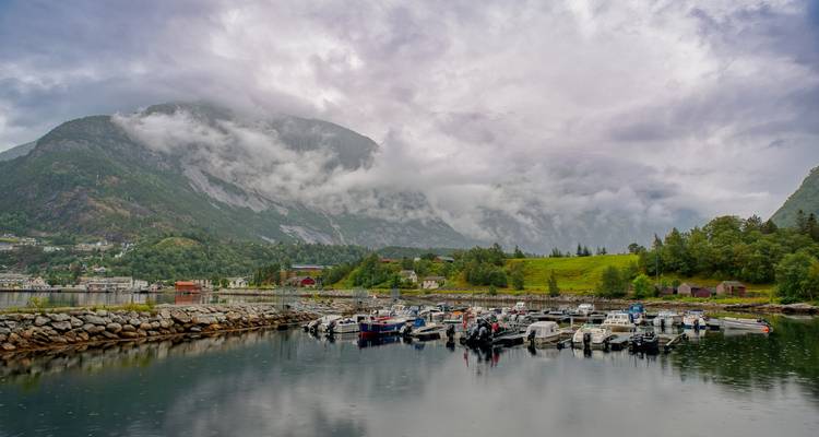 Marina tranquila de pequeñas lanchas motoras bajo montañas envueltas en niebla y un fiordo majestuoso.