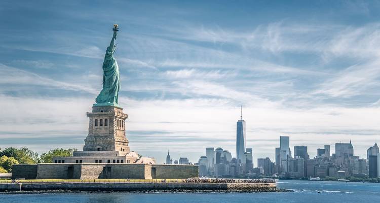 Statue de la Liberté se dressant devant la skyline de Lower Manhattan sous un ciel légèrement nuageux.