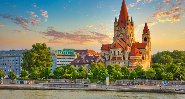 Picturesque riverside scene at sunset with the red-topped towers of St. Francis of Assisi Church in Vienna glowing in warm light beside the Danube.