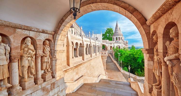Stone arcade walkway inside Fisherman’s Bastion framing a view toward its conical tower and the Danube beyond on a bright morning.