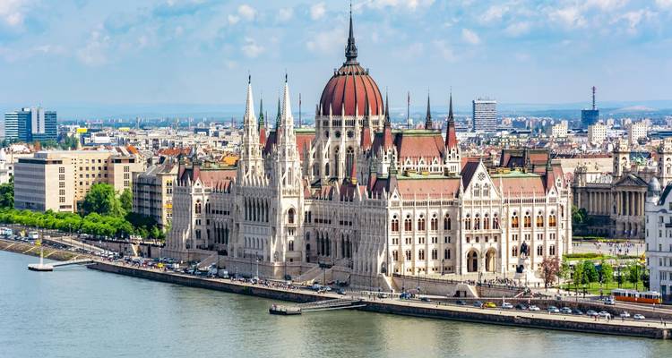 Daytime riverside panorama of the Hungarian Parliament Building rising majestically along the Danube with city skyline in the background.