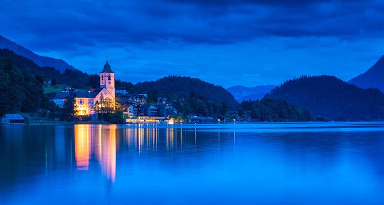 Escena de la hora azul con una pequeña iglesia iluminada y un pueblo reflejados en las aguas tranquilas del lago, rodeados de colinas oscuras.