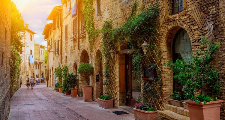 Ruelle étroite et ensoleillée dans un village toscan bordée de maisons en pierre, de vignes grimpantes et de pots en terre cuite, avec quelques piétons au loin.