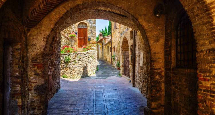 Vue à travers une arche en pierre ombrée révélant une ruelle colorée d'une ville toscane médiévale avec des fleurs et des murs rustiques.