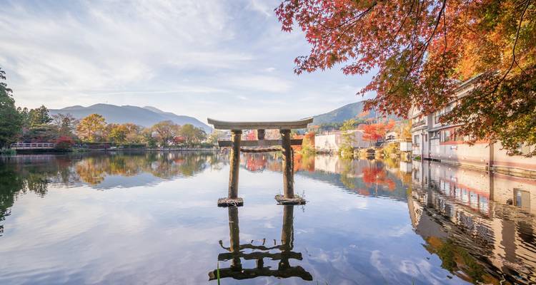 Small torii gate standing in a reflective lake surrounded by colorful autumn trees and mountains.