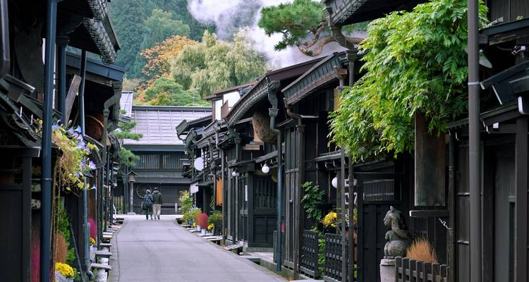 Traditional Edo-period street in Takayama with wooden houses, lush plants and light steam rising in the distance.