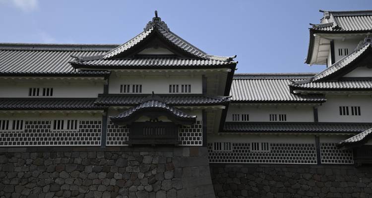 Close view of grey-tiled roofs and white walls of Kanazawa Castle under a pale blue sky.