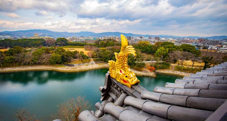 Golden shachihoko ornament on a tiled castle roof overlooks a serene river and sprawling cityscape.