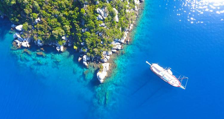 Drone shot of solitary sailboat anchored in crystal-clear blue sea beside lush forested island coastline