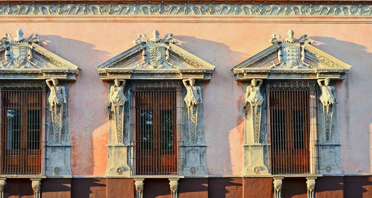 Close-up of ornate colonial façade in warm sunlight with sculpted figures flanking barred windows.