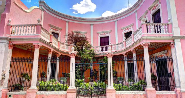 Elegant pink colonial mansion with ornate balconies and wrought-iron fence set against a bright blue sky.