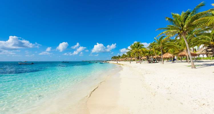 Tropical white-sand beach with turquoise water, palm trees and a few small boats on a clear sunny day.