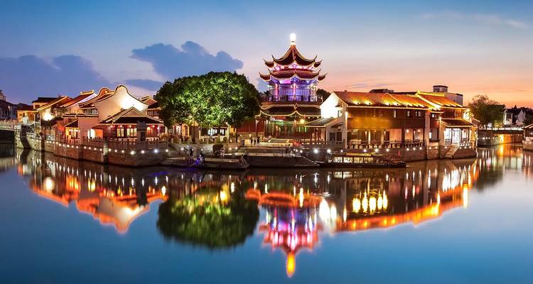 Evening river town with pavilions lit in vibrant colours reflected in the water.