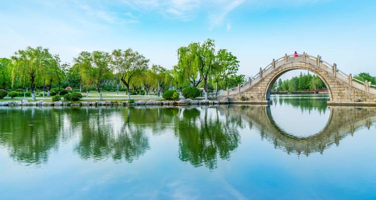 Stone moon bridge mirrored in still lake inside a landscaped Chinese garden.