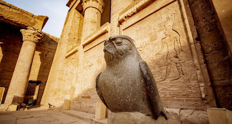 Eine Granitfalkenstatue des Horus steht vor sonnenbeschienenen Säulen und geschnitzten Reliefs im Edfu-Tempel.