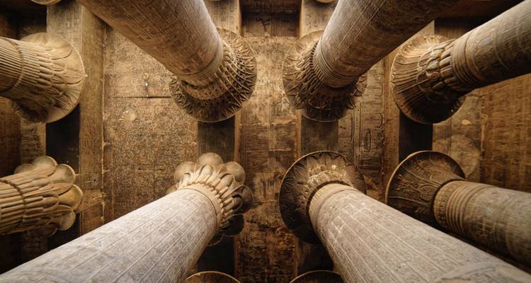 Aufwärtsblick auf kunstvoll geschnitzte Papyrussäulen in einem antiken Tempel, deren Muster durch warme innere Beleuchtung hervorgehoben werden.