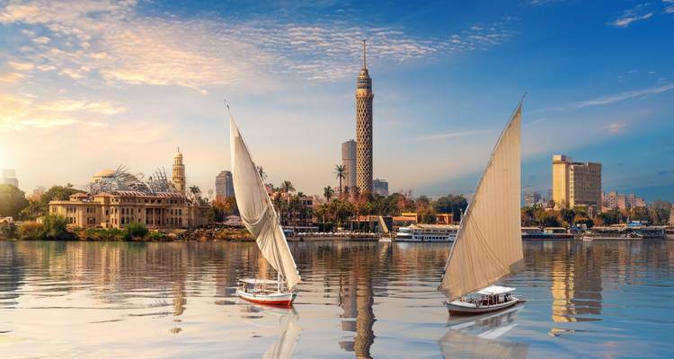 Zwei Felucken segeln friedlich bei Sonnenaufgang auf dem Nil mit dem Cairo Tower und der Skyline der Stadt im goldenen Licht.