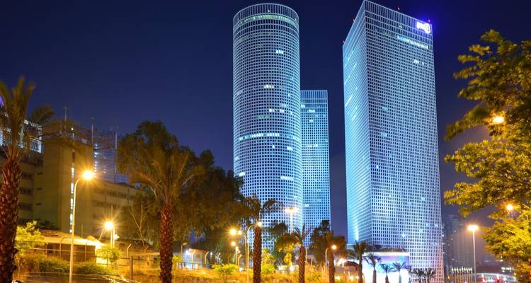 Nachticht van Tel Avivs verlichte Azrieli Center wolkenkrabbers omlijst door palmbomen.