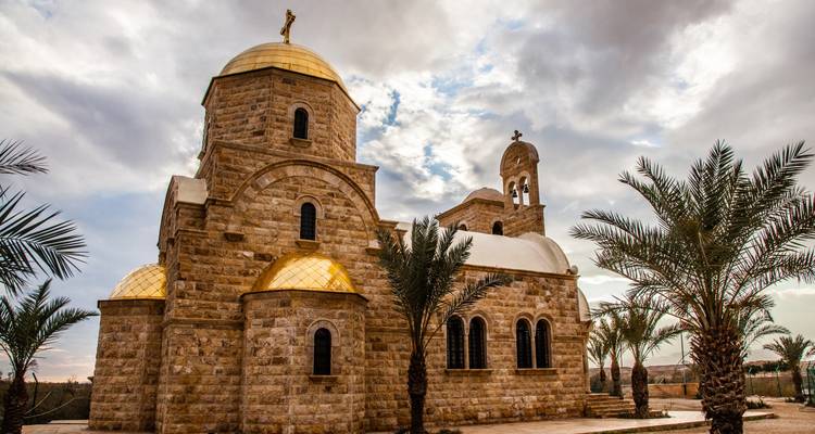 Une église en pierre aux dômes dorés et aux palmiers se dresse sous des ciels nuageux dramatiques.