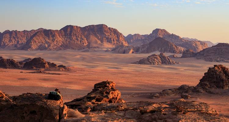 Les vastes montagnes de roches rouges de Wadi Rum brillent dans une lumière douce tandis qu'un randonneur solitaire se repose sur un rocher dominant le désert.