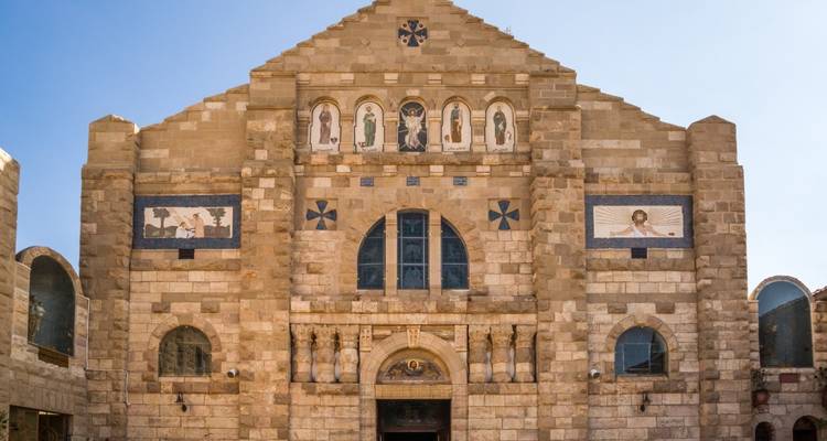 Façade d'église en pierre ornée de mosaïques et de fenêtres en arc contre un ciel sans nuages.