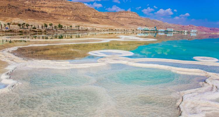 Intricate salt patterns and turquoise water along the shore of the Dead Sea with barren mountains beyond.