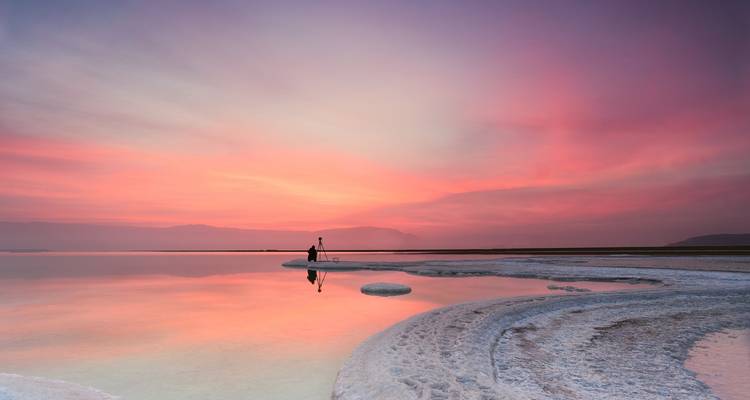 Lever de soleil pastel paisible sur la Mer Morte avec une personne solitaire se tenant sur des formations de sel reflétées dans une eau calme.