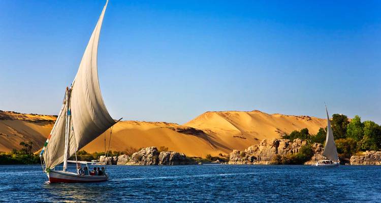 Traditionelles Felucca-Segelboot, das am Nil in der Nähe von goldenen Sanddünen und Granitblöcken gleitet.
