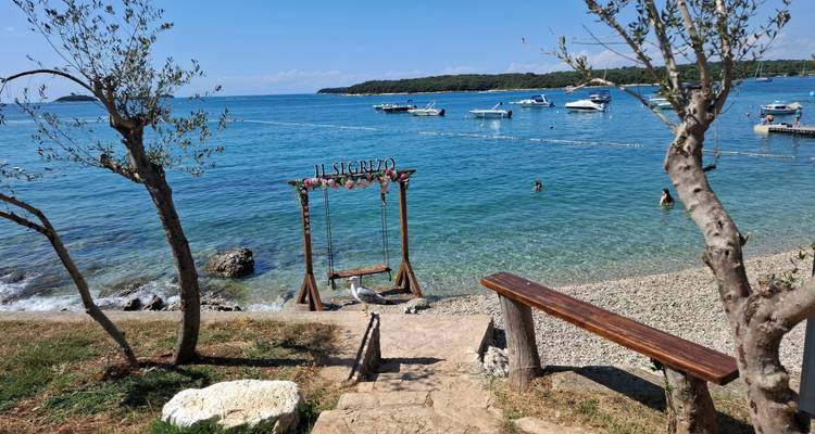 Des marches de pierre mènent à un petit portique de balançoire sur une plage de galets avec des baigneurs et des bateaux amarrés dans une eau bleu vif.