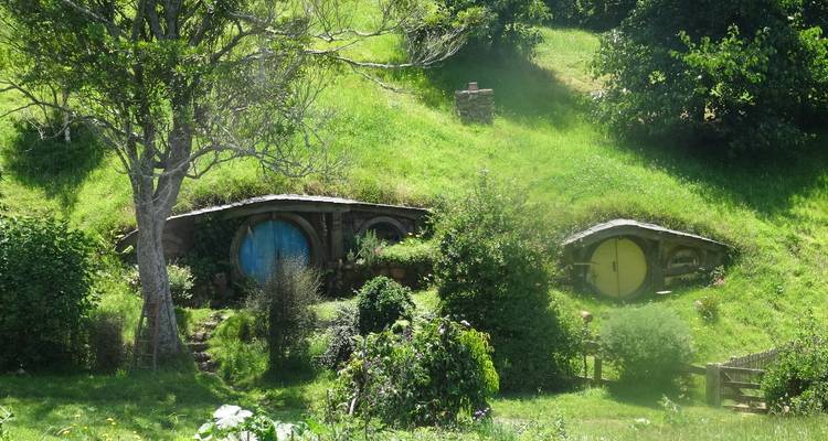 Two neighboring hobbit holes with blue and yellow doors nestled in bright green hillside.