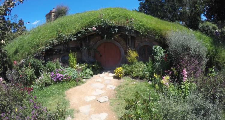 Flower-filled entrance to a hobbit hole with red door set in a grassy mound.